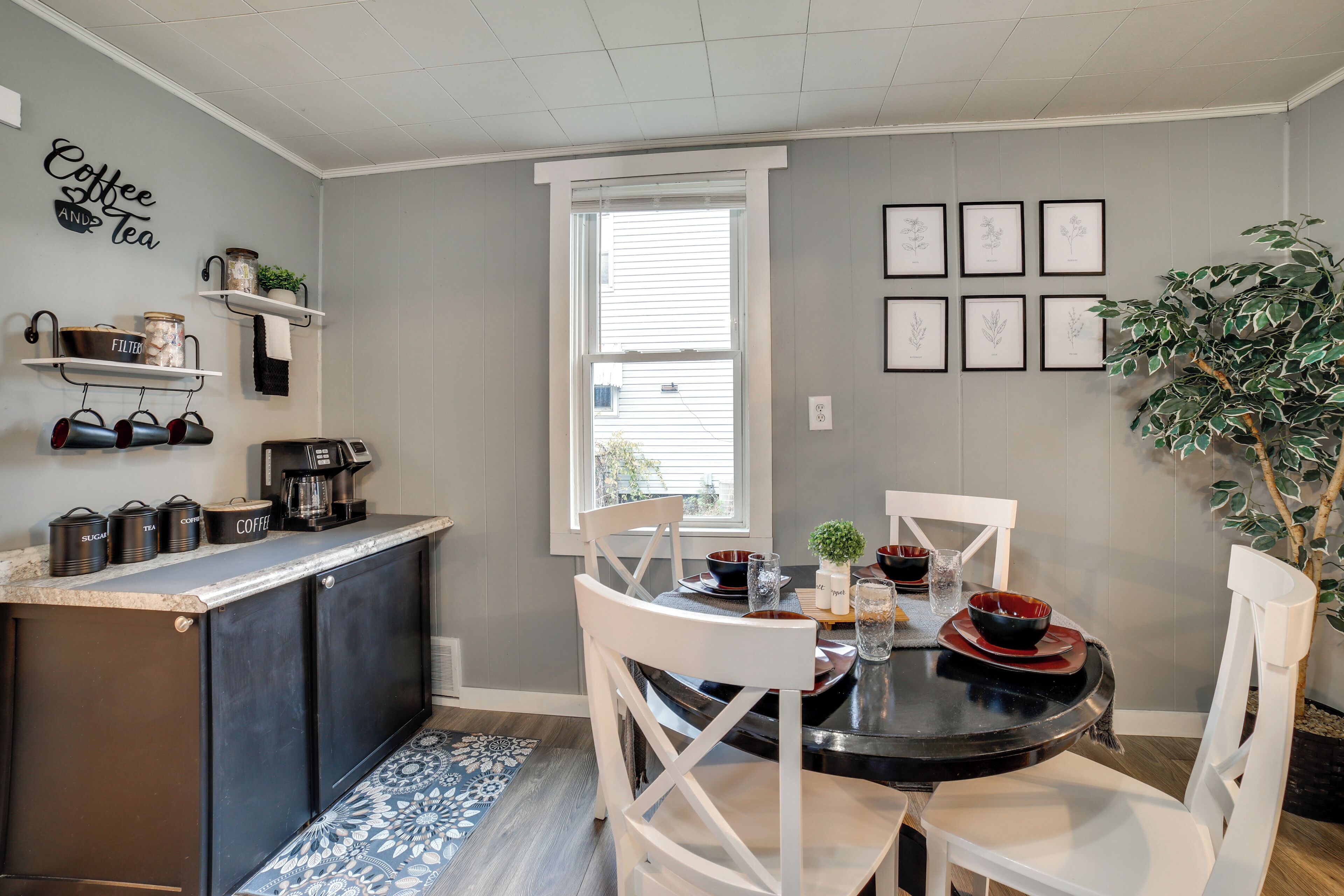A modern kitchen with a dining area, featuring a window, shelves with kitchen items, and framed botanical illustrations.