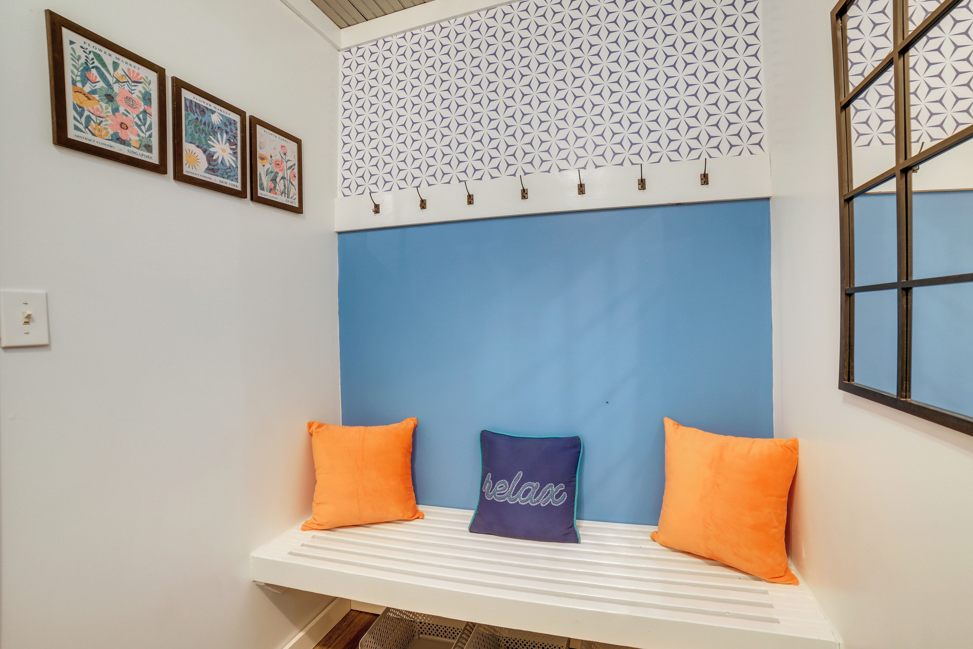 Una habitación con una pared azul de acento, un banco blanco con un cojín naranja y uno morado, y tres obras de arte enmarcadas sobre el banco.