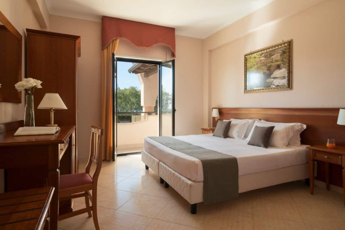 A hotel room with a bed, a desk, a chair, a balcony with a view, and a painting on the wall.