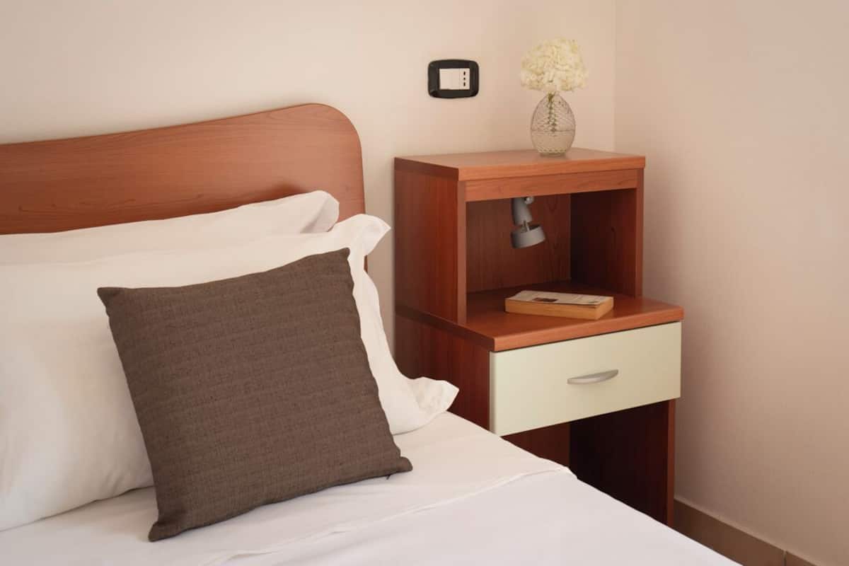 A neatly made bed with white and brown pillows, a wooden nightstand with a drawer, and a vase with white flowers.