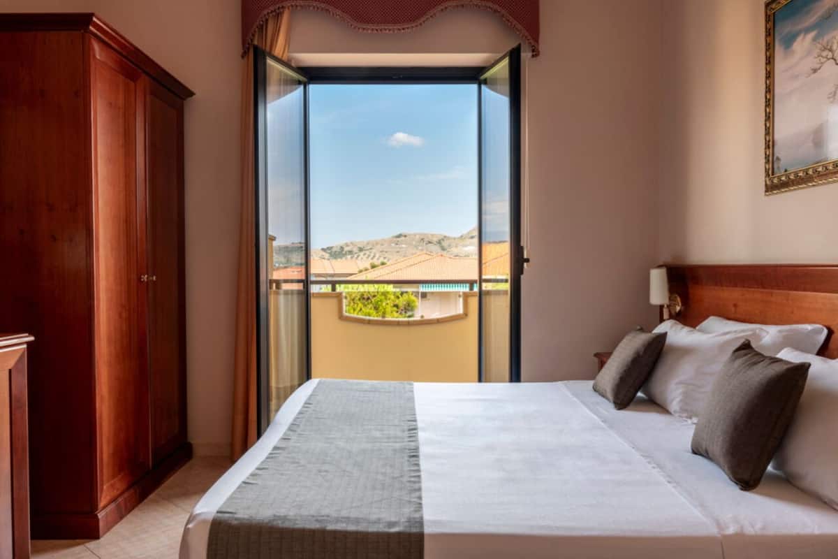 A hotel room with a large bed, a wardrobe, and a balcony with a view of a desert landscape.