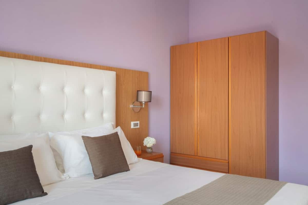 A bedroom with a large bed, a wooden wardrobe, and a nightstand with a lamp and vase.