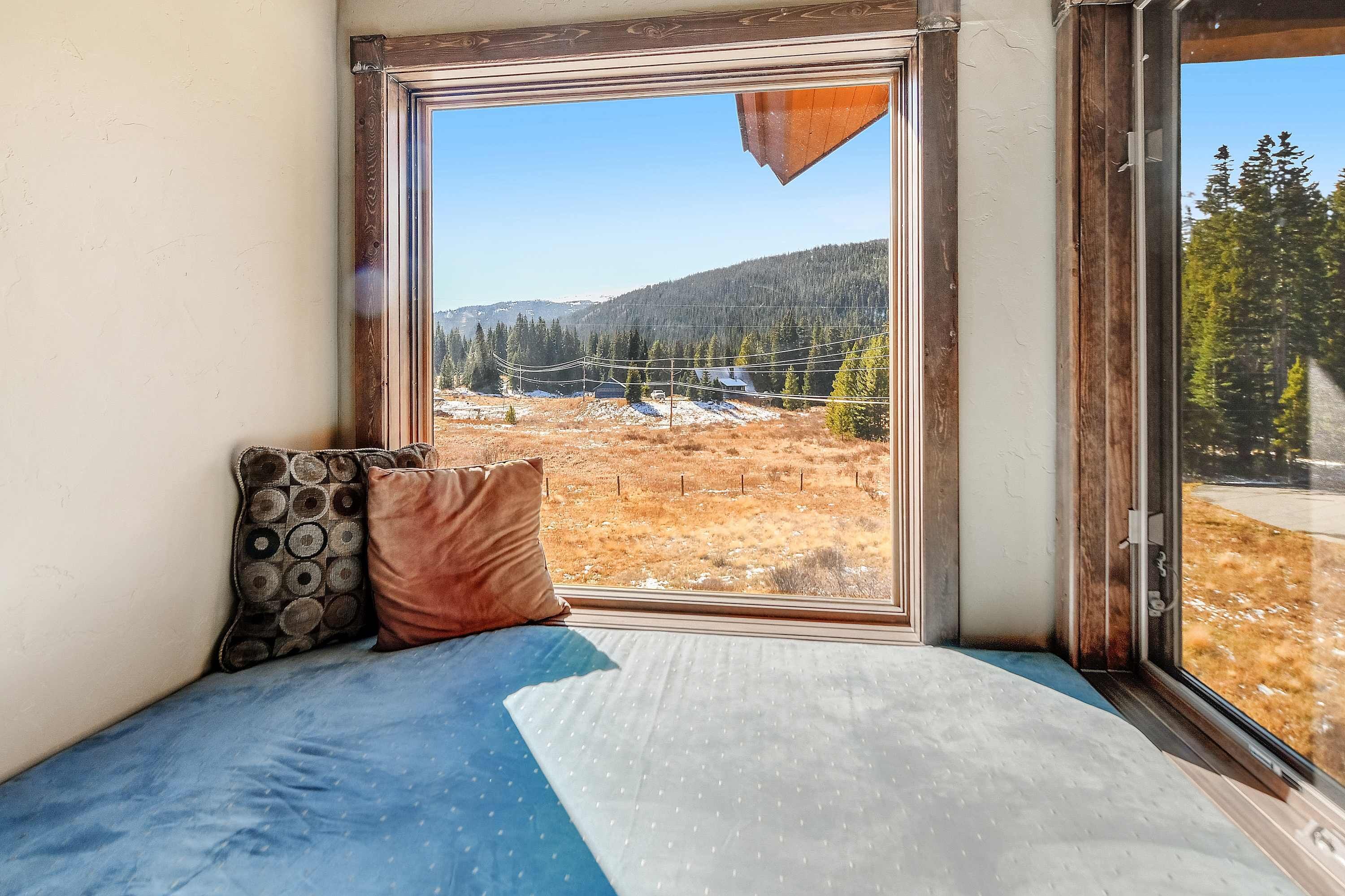 A room with a large window, a bed with patterned pillows, and a view of a forested landscape.