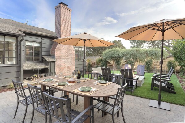 Un patio al aire libre con una mesa de comedor preparada para cuatro personas, rodeada de sillas y dos sombrillas grandes.