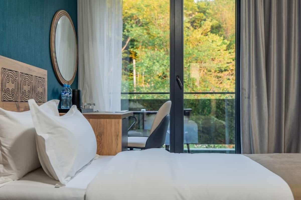 A hotel room with a bed, a desk with a chair, a large window with a view of trees, and a mirror on the wall.