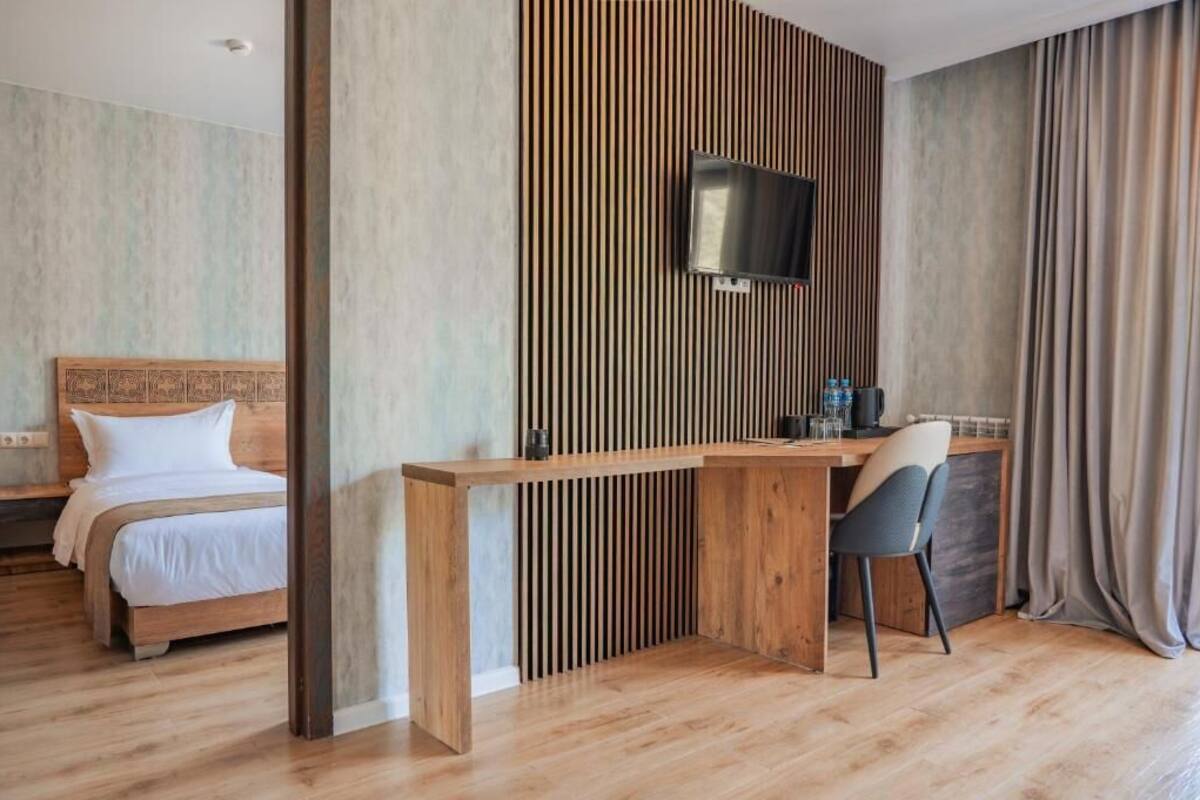 A hotel room with a wooden desk, a television mounted on a slatted wall, a bed with white linens, and a curtain on the window.