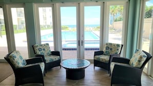 Ein sonnendurchfluteter Innenbereich mit Korbmöbeln und einem gefliesten Couchtisch, mit Blick auf einen Swimmingpool und eine Wohngegend.