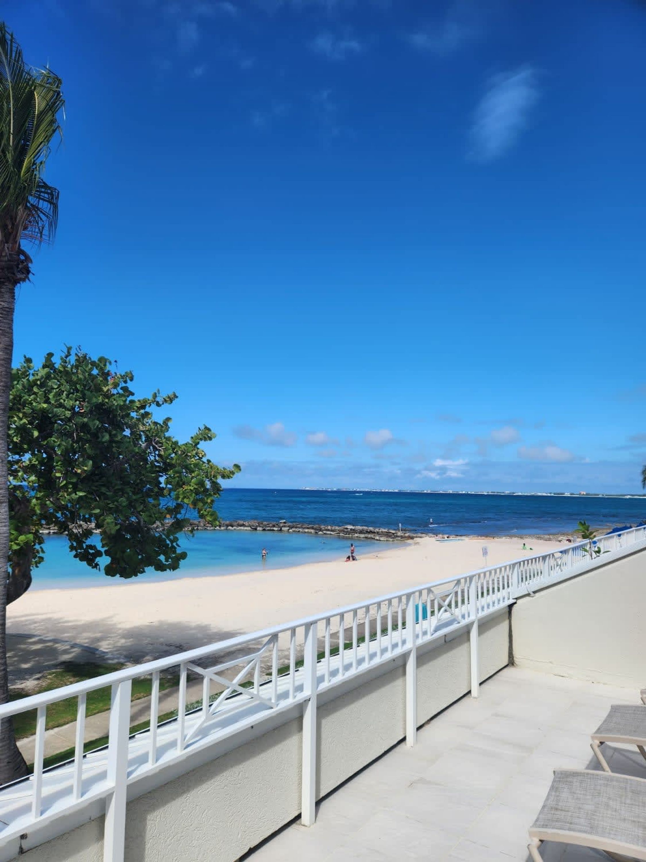 A beach view from a balcony with a white railing, clear blue sky, and calm sea.