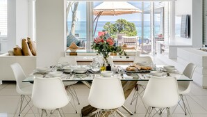 A modern dining area with a glass table, white chairs, and a view of the sea. - Villa Bleu Blanc in Camps Bay With Pool and Views (Cape Town)