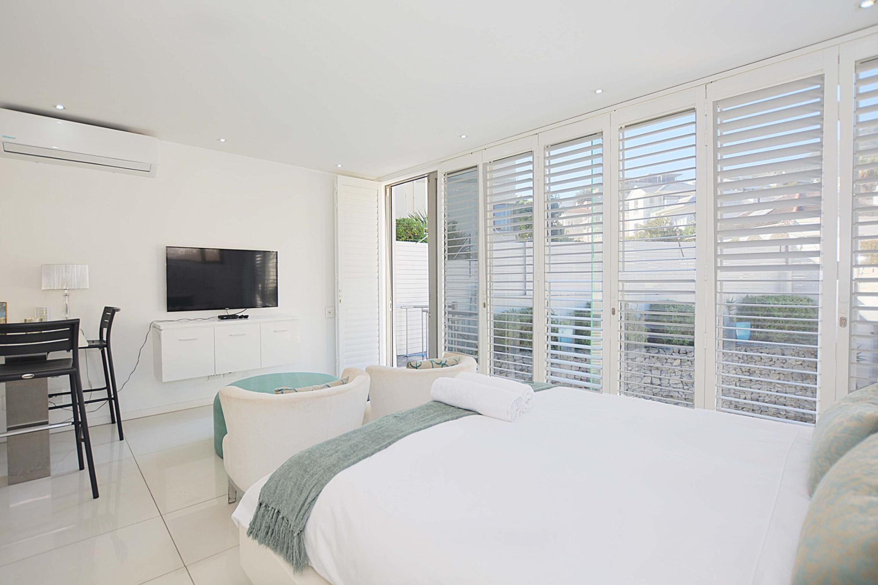 A modern bedroom with a large bed, a desk with a chair, a flat-screen TV, and a balcony with white shutters.