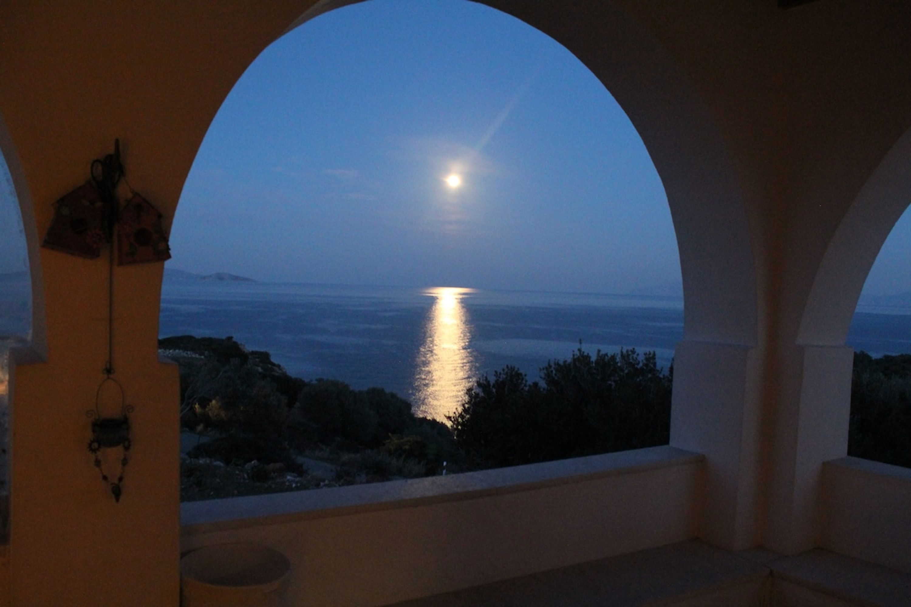A view through an arched window overlooking a body of water at night with a moon visible in the sky.
