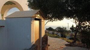A stone pathway leading to a small white building with a sloped roof, surrounded by trees and a view of the sea. - Villa Rouvis With Outdoor Hot Tub (Naxos)