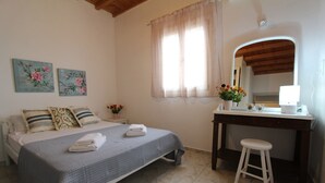 A bedroom with a bed, a desk, a chair, a mirror, and two floral paintings on the wall. - Villa Rouvis With Outdoor Hot Tub (Naxos)