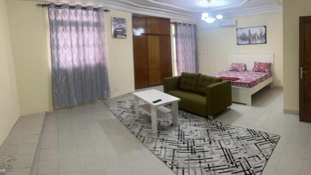 Una habitación con un sofá verde, una mesa de centro blanca, una alfombra estampada, una cama con ropa de cama rosa y un cuadro de un paisaje urbano en la pared.