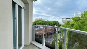 Un balcón con barandilla de vidrio, una puerta corrediza de vidrio y vista a una zona residencial con árboles y edificios.