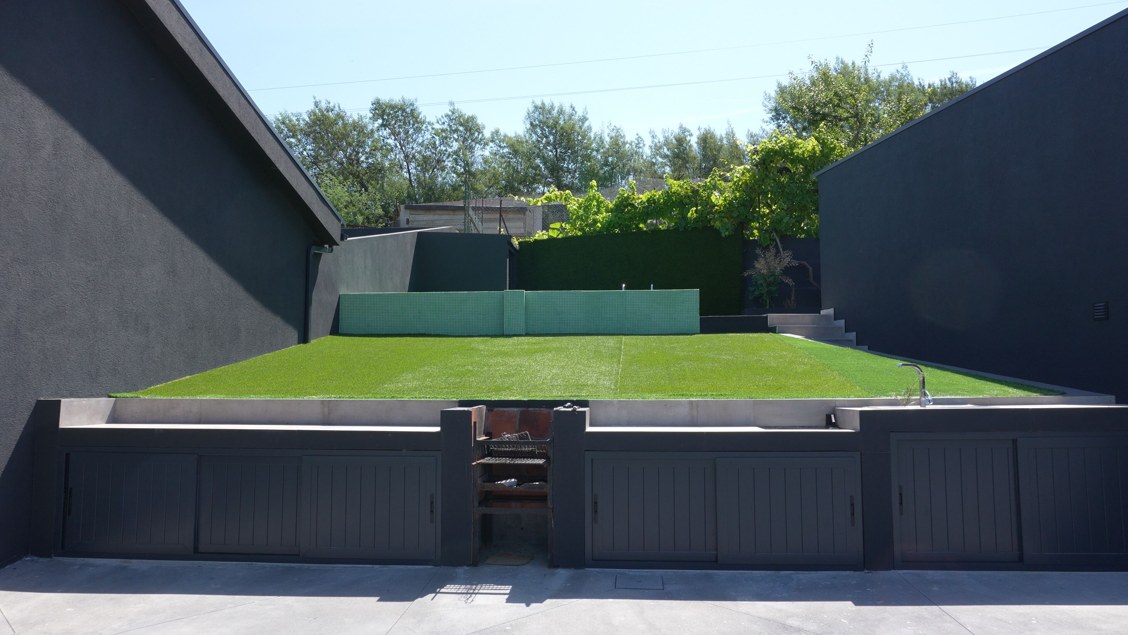 Un espacio exterior moderno con césped plano, valla verde y una parrilla de metal.