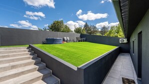 Un espace extérieur moderne avec une pelouse artificielle verte, des marches menant à une plateforme surélevée et un mur noir.