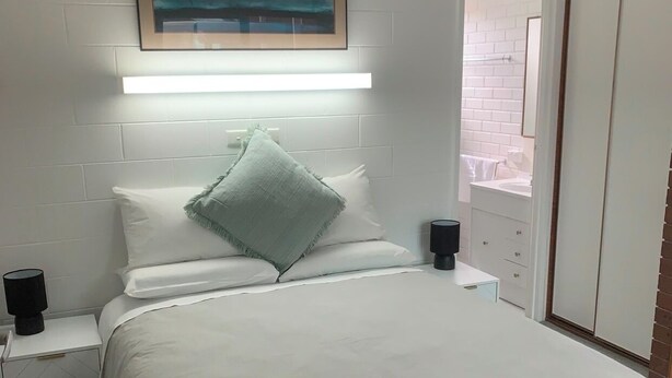 Een slaapkamer met een groot bed, witte beddengoed, een schilderij boven het bed en een nachtkastje met een lamp.