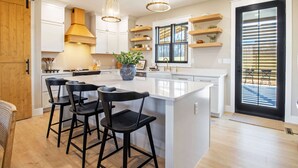 A modern kitchen with a central island, black bar stools, pendant lights, and wooden accents.