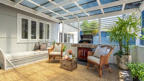 Een overdekt terras met rieten meubels, een barbecue en plantenpotten.