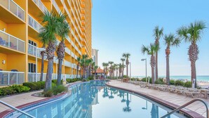 Un espace piscine d’hôtel offrant une vue dégagée sur l’océan, entouré de palmiers et bordé d’un bâtiment jaune doté de balcons.