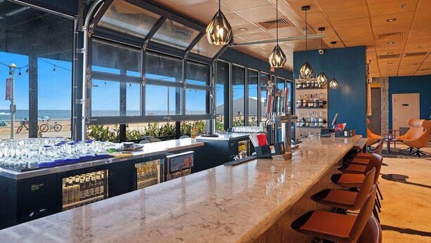 A bar area with a long counter, bar stools, and a view of the beach. - Boardwalk Below, Beach Ahead (Virginia Beach)