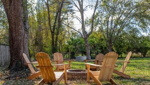 Disposición circular de asientos con cuatro sillas Adirondack de madera alrededor de un brasero central, ubicada en un patio trasero arbolado con árboles y una valla de madera al fondo.