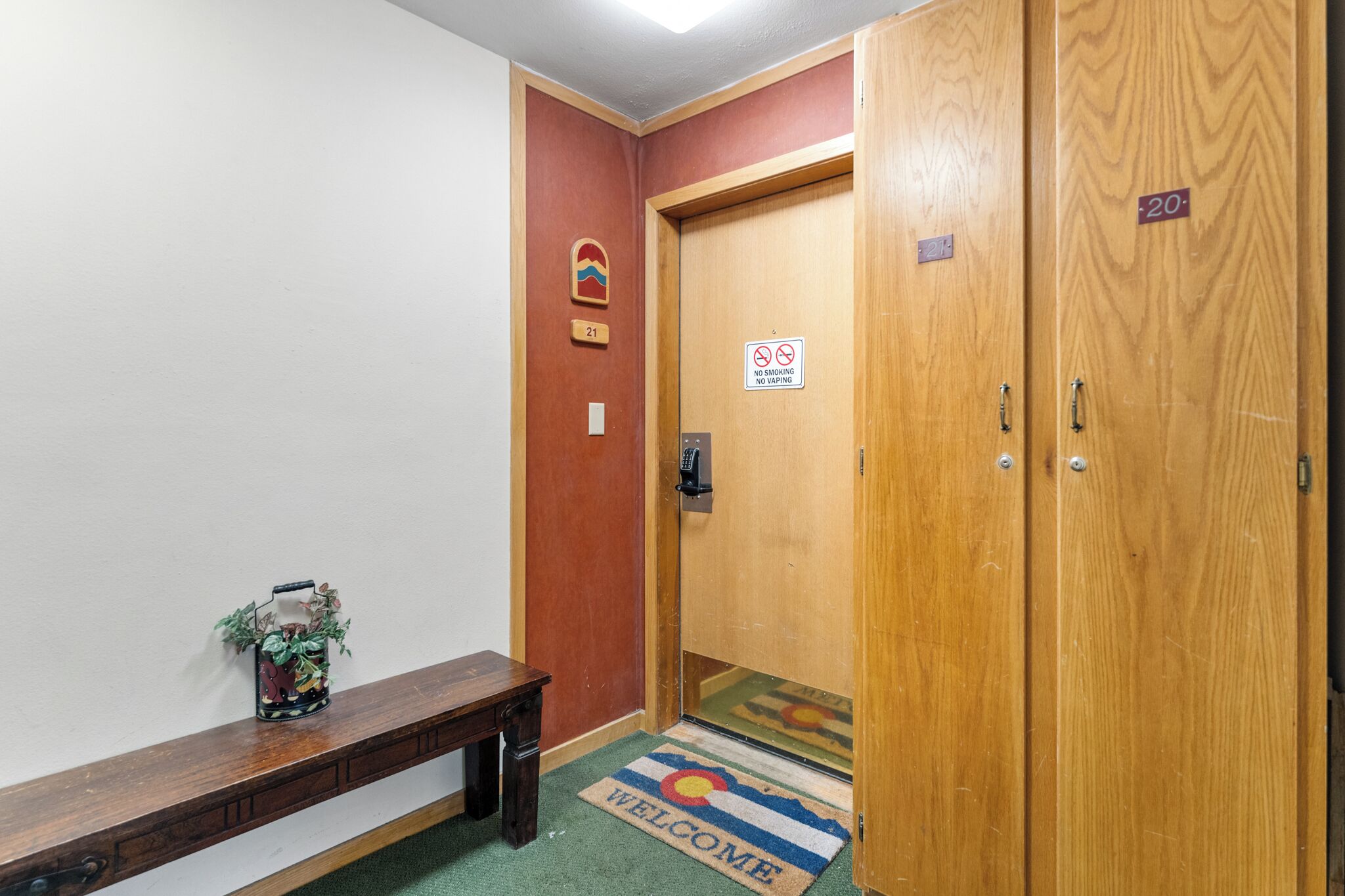 Un couloir d’hôtel avec des portes en bois, un banc, un paillasson et un panneau indiquant qu’il est interdit de fumer.