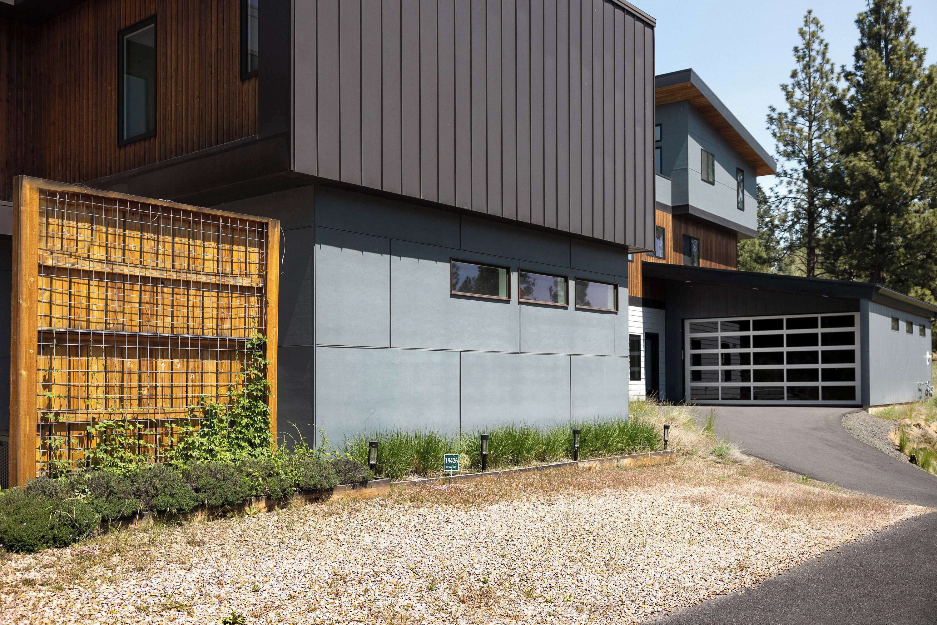 A modern two-story house with a garage, a gravel driveway, and a wooden fence with climbing plants.