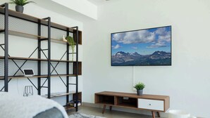 En moderne stue med et fjernsyn ophængt på væggen, et træ konsolbord og en reol med en bærbar computer og planter.