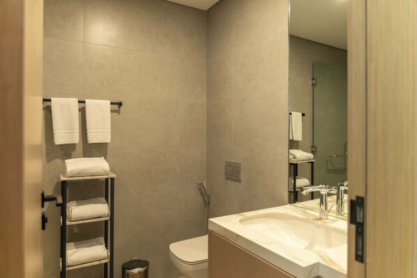 A modern bathroom with a sink, toilet, and shower. There are towels on a rack and a shelf. - Pearlhouse Bahrain (Manama)