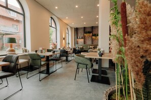 A modern cafe with tables and chairs, large windows, and indoor plants.