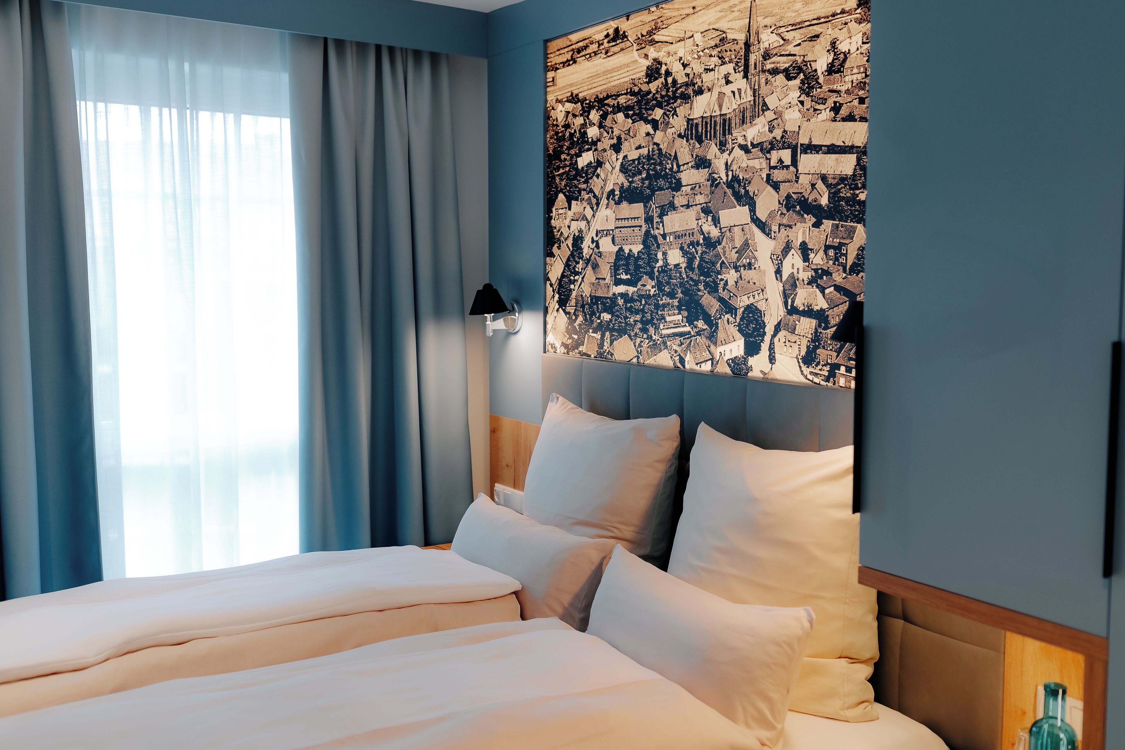 A hotel room with a large bed, a nightstand, a lamp, and a cityscape artwork on the wall.