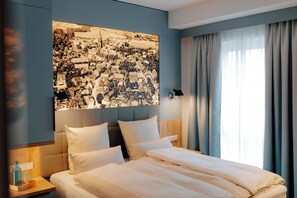 A hotel room with a large bed, a nightstand with a lamp, and a cityscape artwork on the wall.