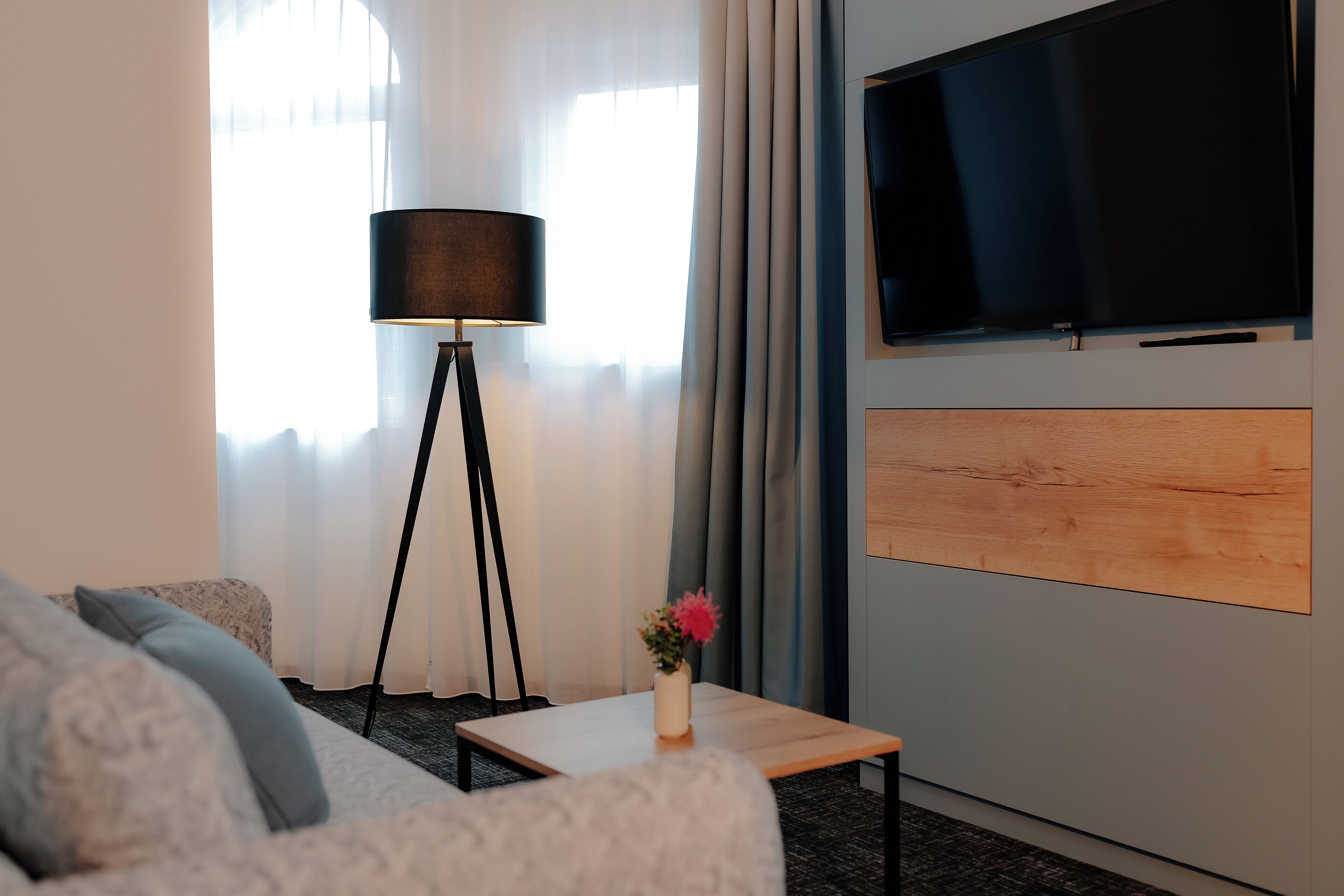 Una habitación de hotel moderna con un sofá, una pequeña mesa con un florero, una lámpara de pie y un televisor de pantalla plana montado en la pared.