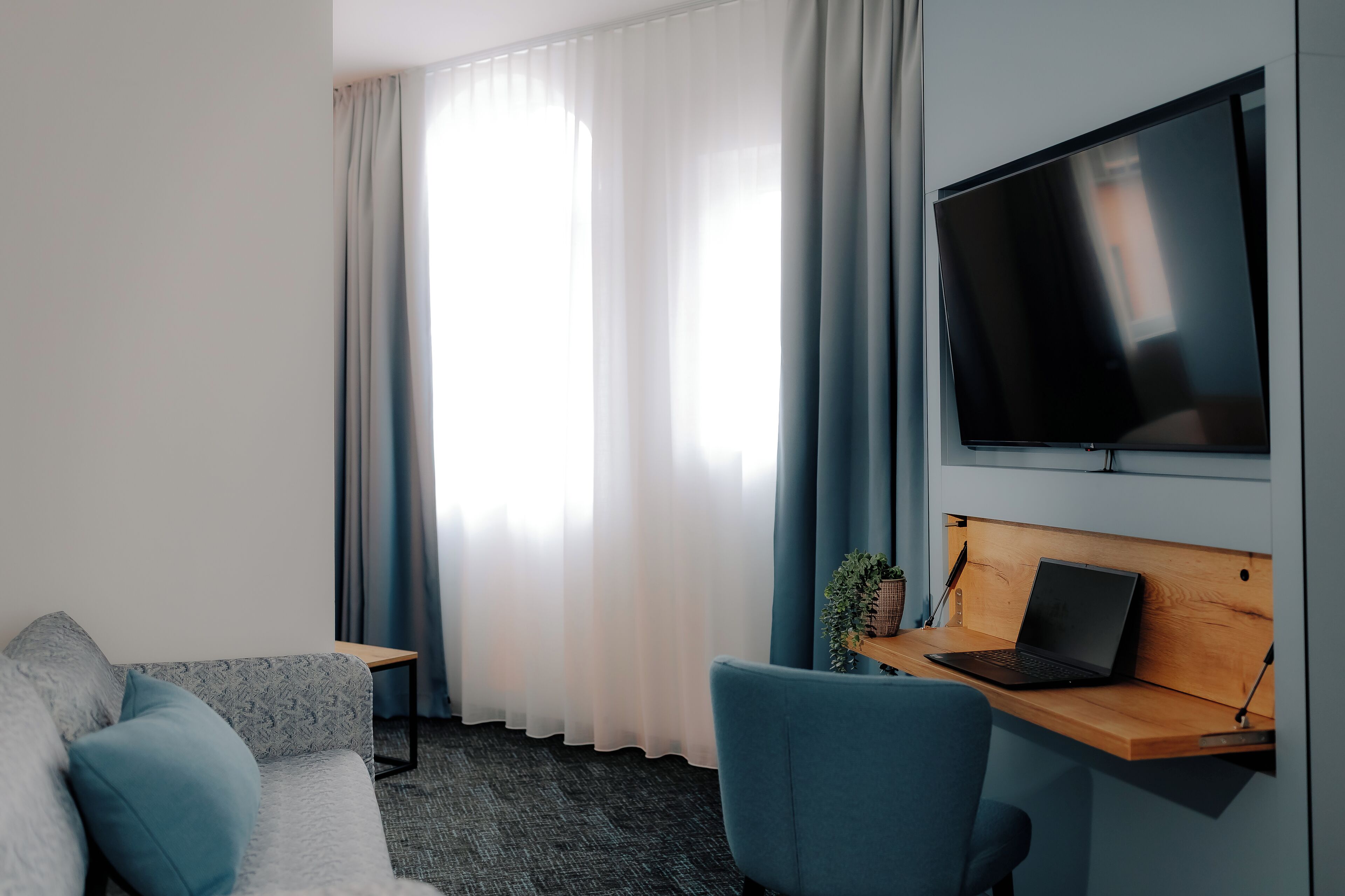 Una habitación de hotel moderna con sofá, escritorio con computadora portátil, televisión y un gran ventanal con cortinas.