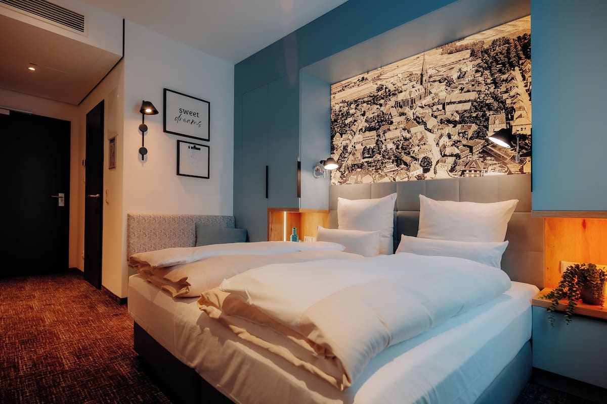 Ein Hotelzimmer mit einem großen Bett, einem Stadtbild-Wandgemälde und einem kleinen Tisch mit einer Lampe.