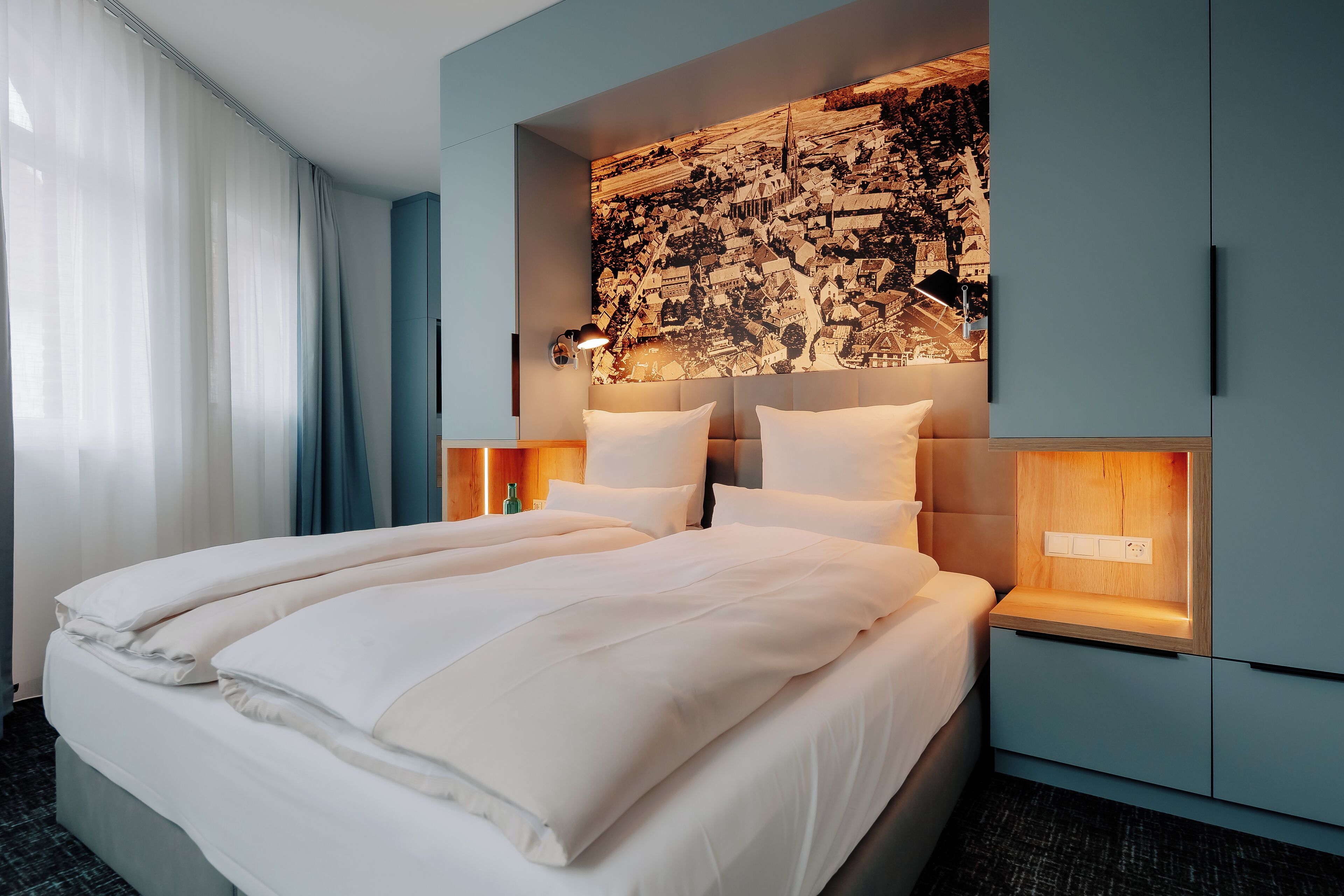 A modern hotel room with a large bed, a cityscape mural on the wall, and built-in bedside tables with lamps.