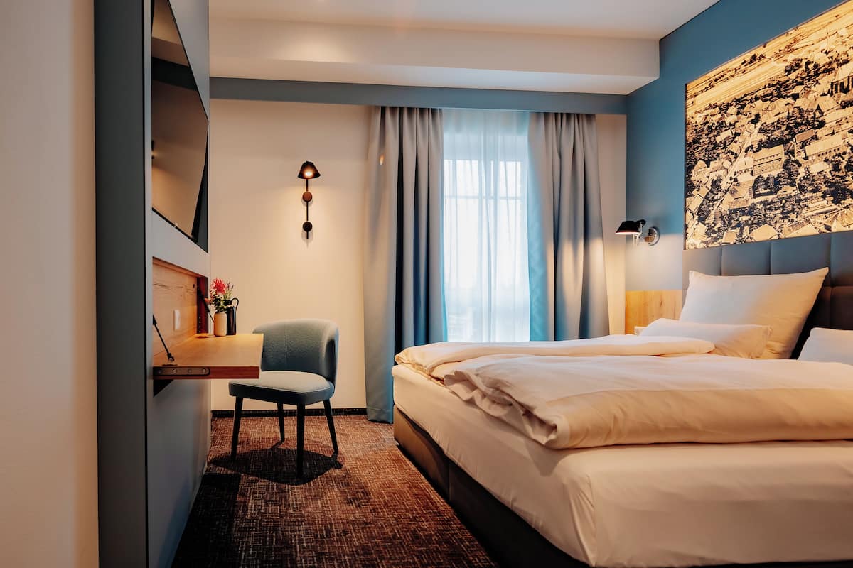 Ein Hotelzimmer mit Bett, Schreibtisch, Stuhl, einer Wandlampe und einem großen Fenster mit Vorhängen.