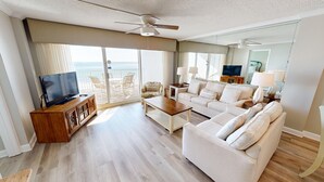 Amplia sala de estar con un sofá grande, una mesita de centro y un televisor sobre un mueble de madera. Cuenta con un balcón con vistas al océano.
