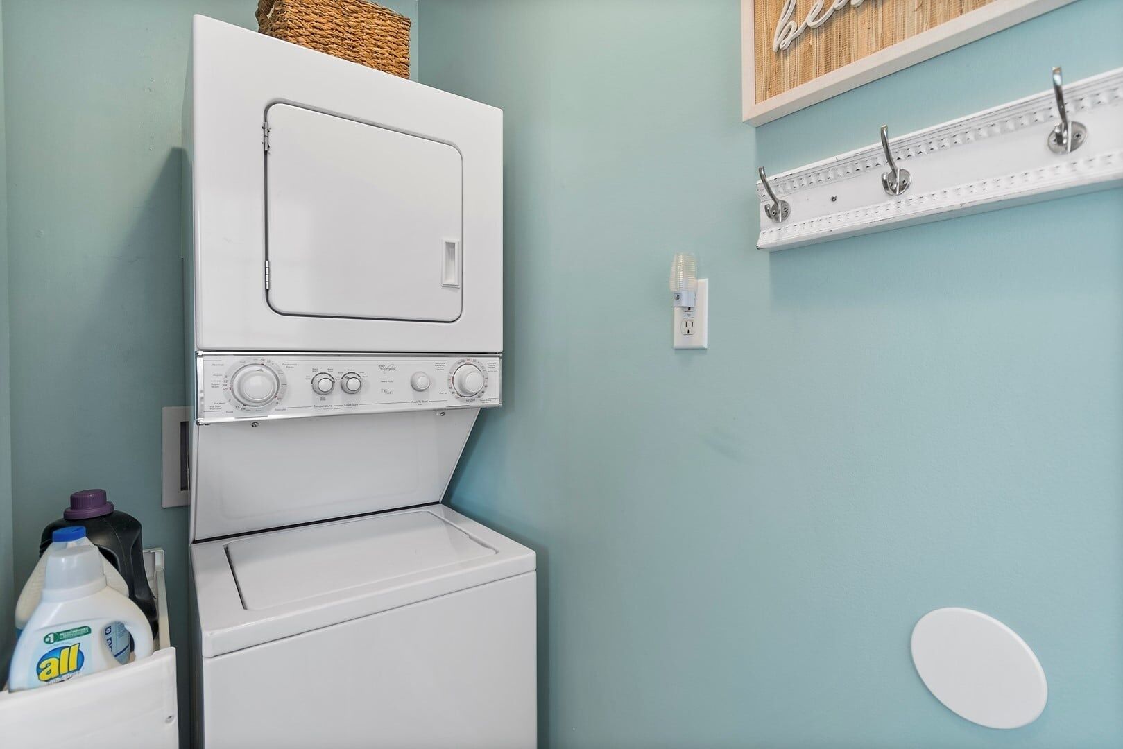 A laundry area with a stacked washer and dryer, a shelf with hooks, and a bottle of detergent.