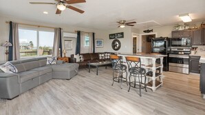 Un salon moderne avec un canapé d’angle, un coin repas doté de tabourets de bar, une cuisine équipée d’appareils électroménagers en acier inoxydable et un ventilateur de plafond.