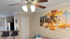 Un salon avec un canapé beige, un tapis bleu, un ventilateur de plafond et une cuisine en arrière-plan.