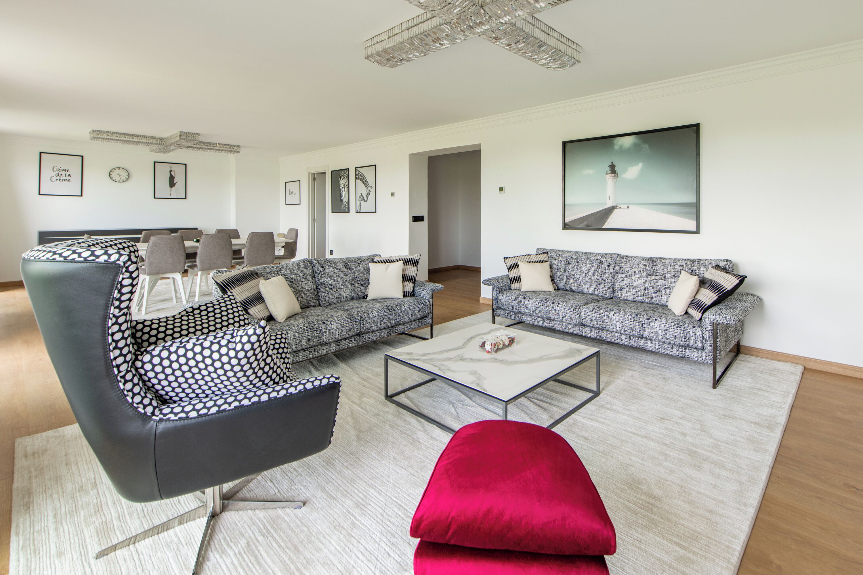 Ein modernes Wohnzimmer mit einem grauen Sofa, einem schwarzen Sessel und einem roten Hocker. Ein Glastisch und ein Gemälde eines Leuchtturms sind an der Wand zu sehen.