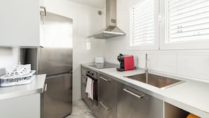 Una cocina moderna con electrodomésticos de acero inoxidable, una encimera blanca y un ventanal con persianas.