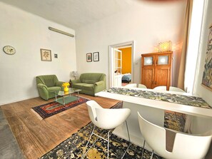 A compact living space with a dining area, two green armchairs, a glass coffee table, a white dining table, and a wooden cabinet.