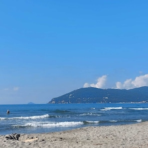 A beach with waves, a clear sky, and a mountain in the background. - Between the Mountains and the Sea (Carrara)
