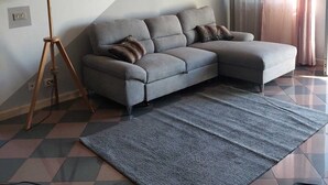 Ein Wohnzimmer mit einem grauen Ecksofa, einer Stehlampe, einem Gemälde und einem großen Fenster mit Vorhängen.