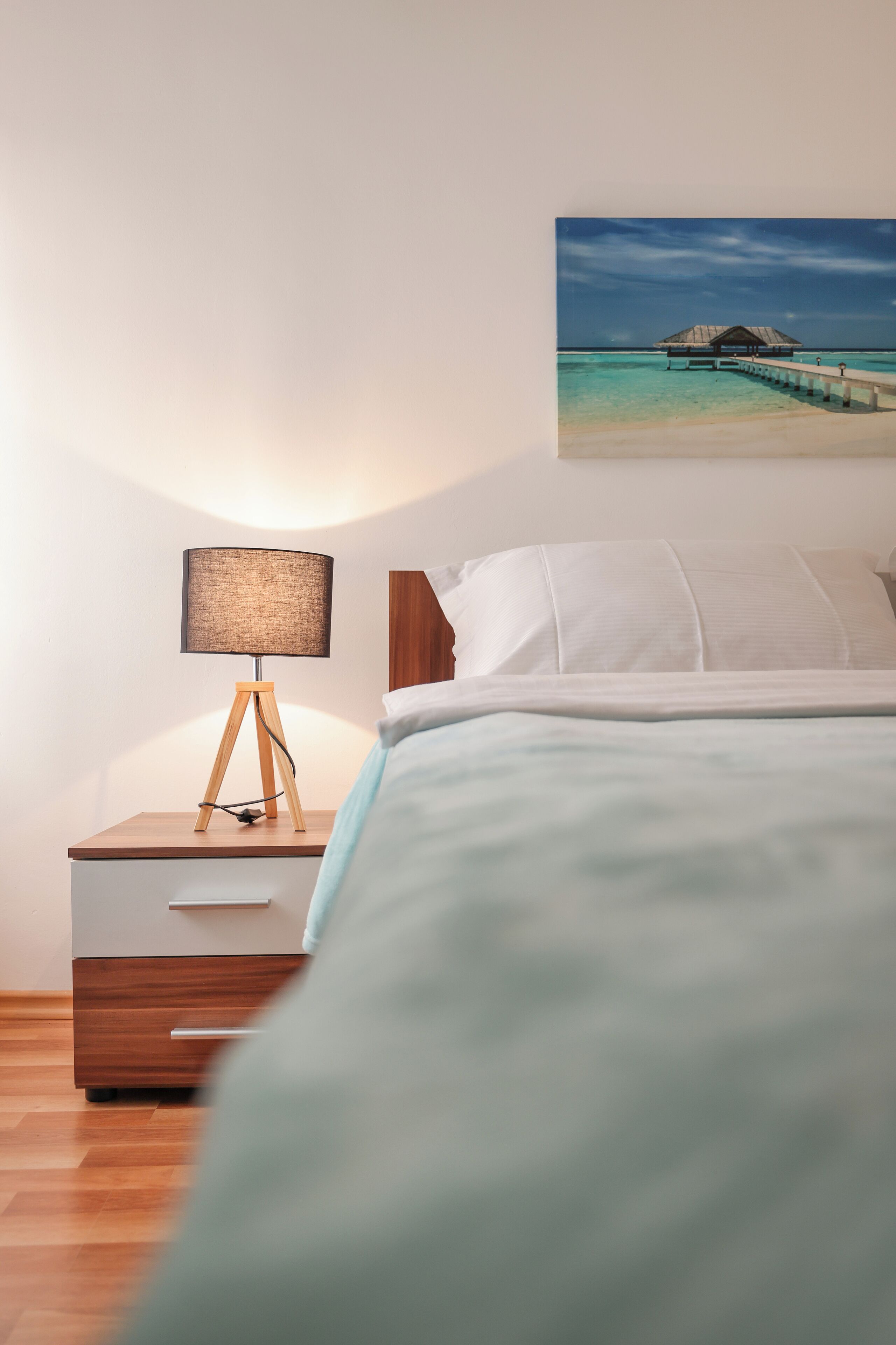 A bedroom with a wooden nightstand, a lamp, a bed with white bedding, and a picture of a beach on the wall.