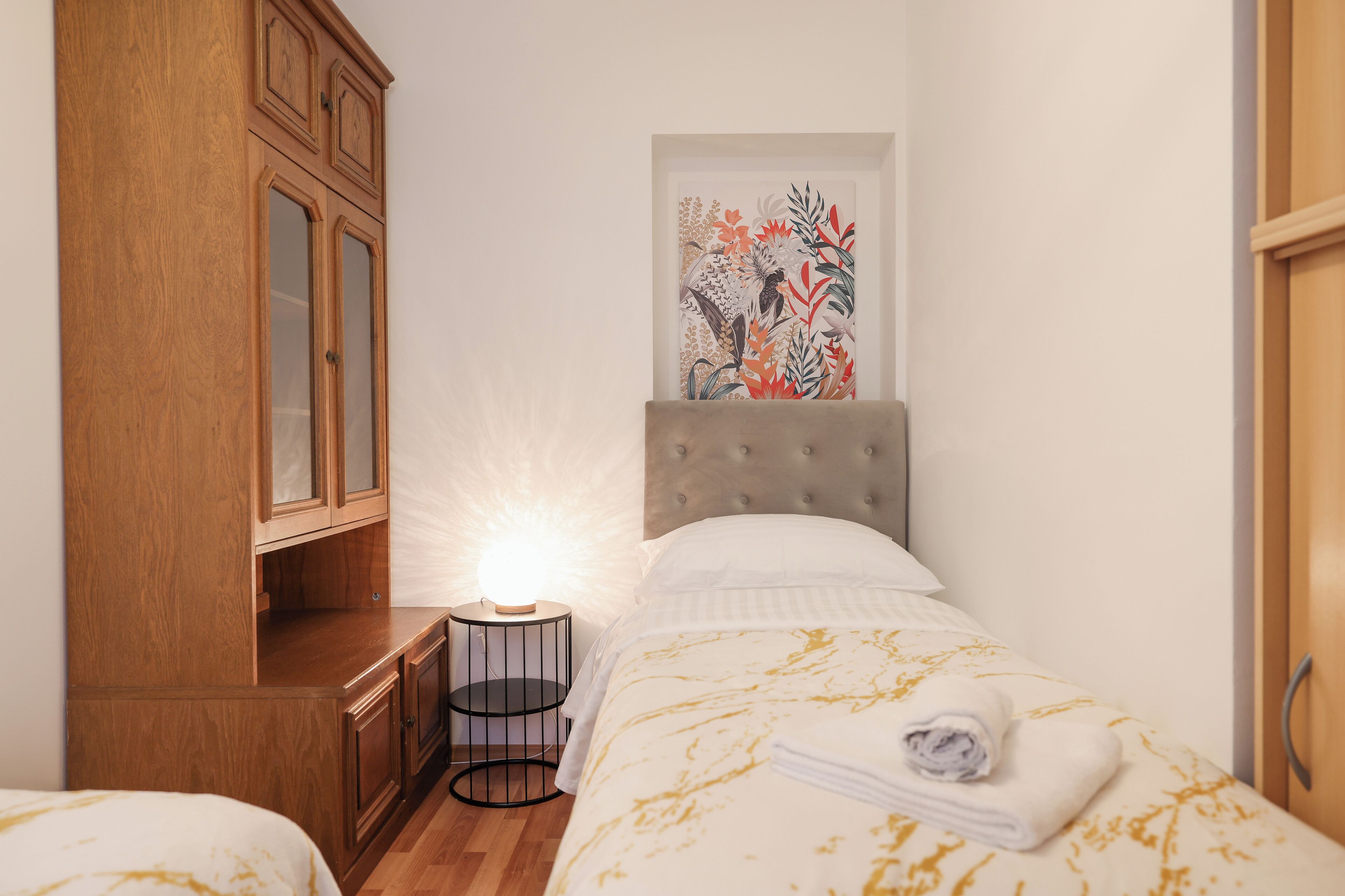 A bedroom with a bed, a nightstand, a wooden wardrobe, and a framed floral artwork on the wall.